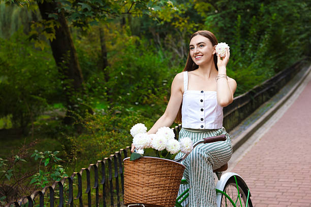 Woman riding bicycle with flowers in basket outdoors. Space for text