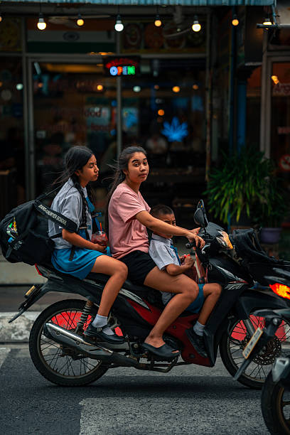 Phuket, Thailand – December 5, 2025: A woman riding a motorcycle with two children as passengers through city traffic.