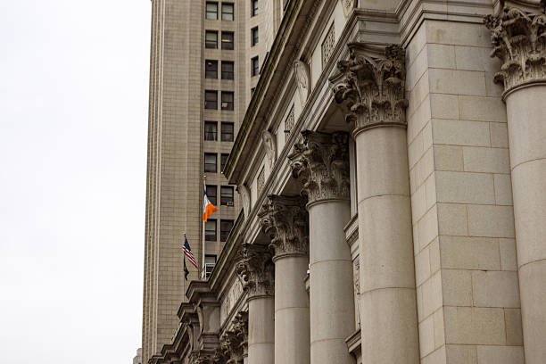 A close-up view of a historical government building featuring large stone columns and detailed architectural elements against a cloudy sky. Flags are visible in the background.
