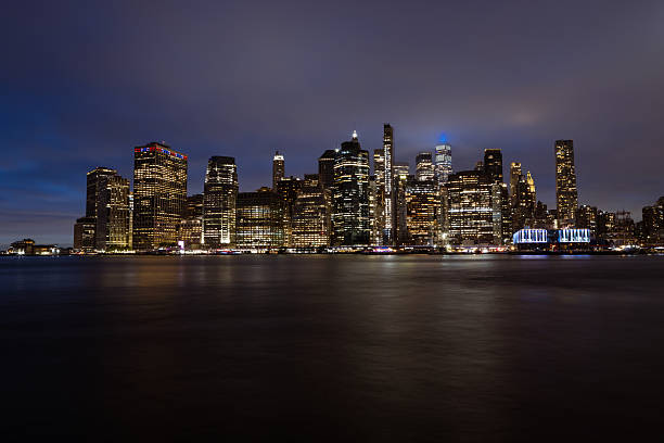 A stunning nighttime view of a city skyline across a calm body of water, showcasing illuminated office buildings and skyscrapers against a dark, cloudy sky.