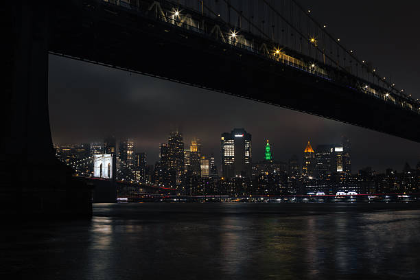 Stunning nighttime view of the New York City skyline with illuminated buildings and a prominent bridge in the foreground, captured from across the river.