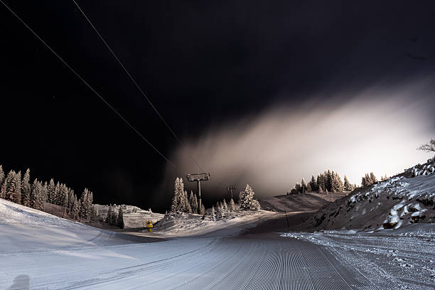 Groomed ski piste glowing under lights at night with snow-covered trees and ski lift, ready for winter sports