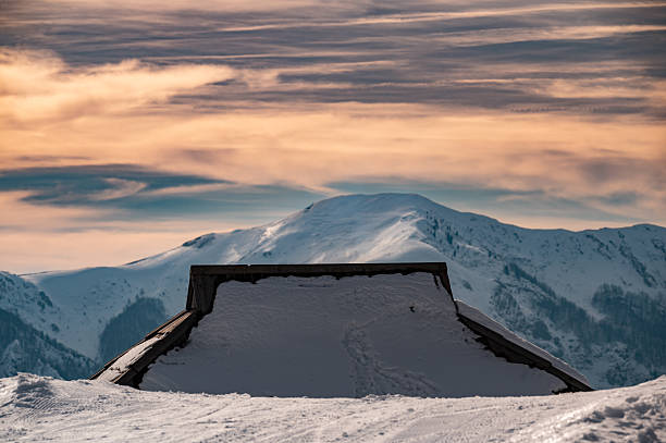 Old mountain hut roof peaking through deep snow on a cold winter day. Warm orange clouds at sunset over snowy peaks