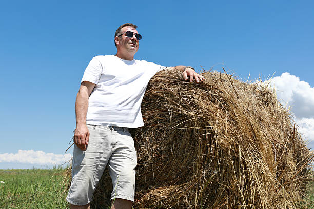 Man in sunglasses leaning on a hay bale in a field under a clear blue sky. Rural summer scene depicting harvest and agriculture.