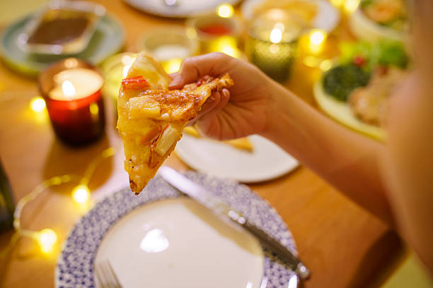 Hand holding slice of pizza during cozy Christmas dinner at home with candlelight and festive decorations. Warm holiday atmosphere, comfort food, casual home dining, and joyful seasonal lifestyle concept.