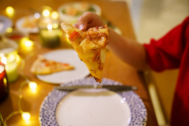Hand holding slice of pizza during cozy Christmas dinner at home with candlelight and festive decorations. Warm holiday atmosphere, comfort food, casual home dining, and joyful seasonal lifestyle concept.