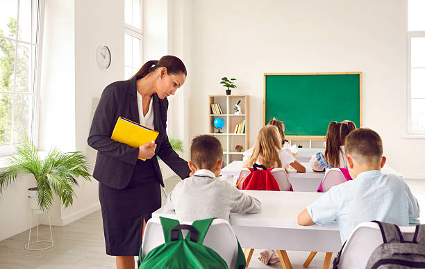 istock School teacher helping her elementary students who are sitting at desks and writing 2253154666