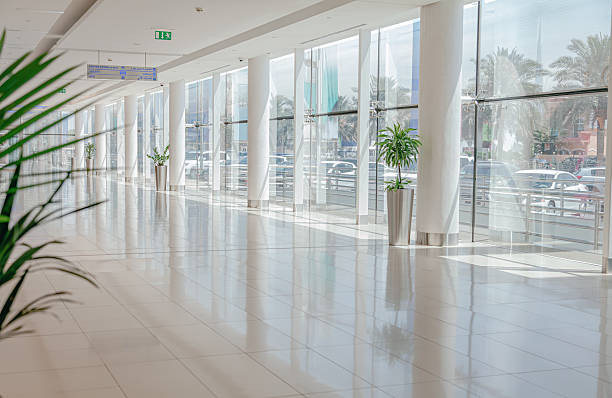 Sunlit empty hallway in a modern shopping mall with clean white tile floor, tall pillars, glass wall, indoor potted palms in metal planters, reflections and long perspective creating a minimalist architectural background with ample copy space for retail, airport, hospital or office concepts
