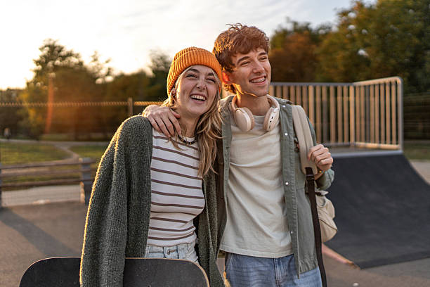 Happy teenage couple standing arm in arm in a skate park at sunset, laughing together while holding a skateboard, sharing a warm, carefree golden-hour moment