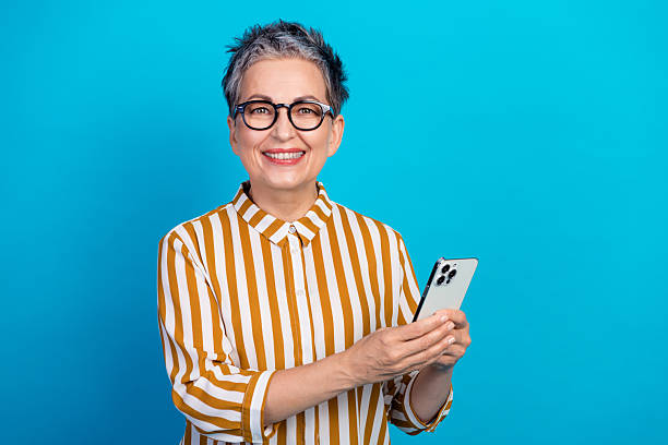 istock Aged female senior smiles while using a smartphone in a striped shirt against a blue background for lifestyle advertising 2253167853