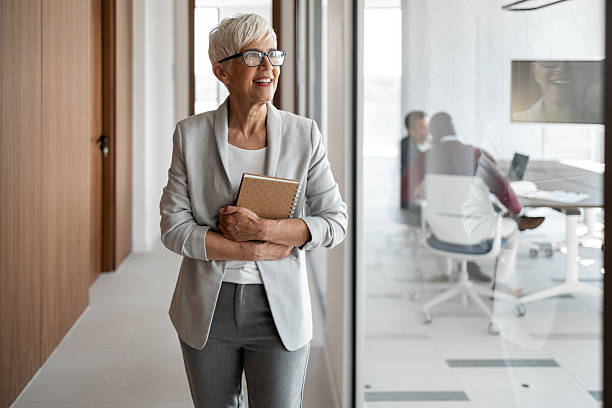 istock Confident Senior Woman In Office Corridor Holding Notebook Smiling While colleagues Work Behind Glass Wall 2253171050