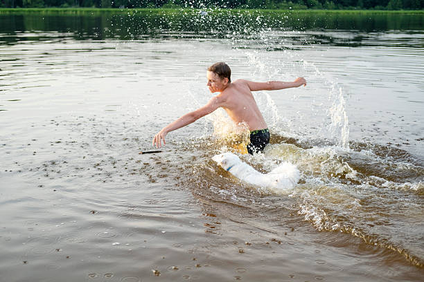 Man and dog play fetch in a lake, causing splashes and excitement. The man is throwing a stick, and the dog eagerly chases it. This image captures dynamic outdoor fun and joyful interaction.