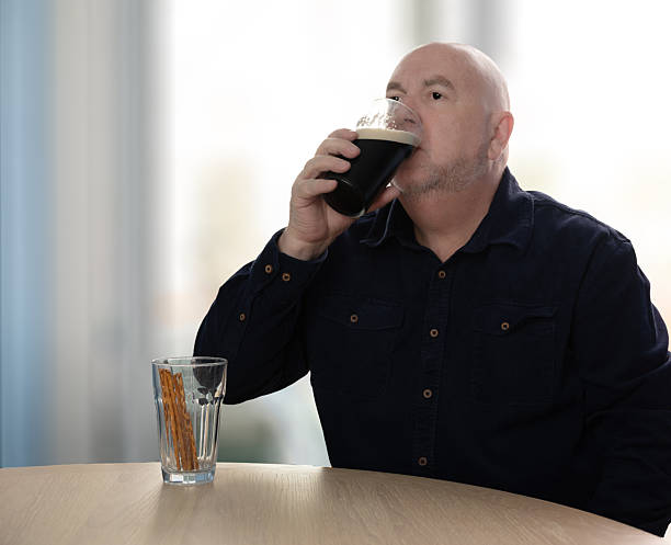 A bald man in a dark blue shirt drinks a dark beer from a glass, with pretzel sticks in a separate glass on the table. New Year’s resolutions, stop drinking