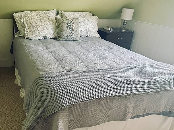 New clean queen bed comforter with slanted ceiling, table, lamp, decorative neutral pillows in bedroom staging model home, house apartment, towels in hostel hotel room