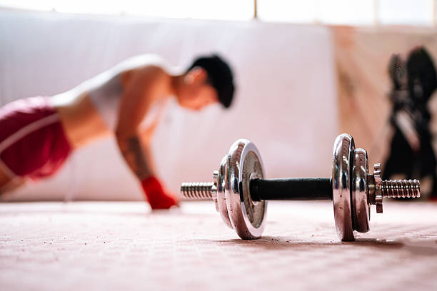 Dumbbell resting on the floor in a gym with an adult person in the background doing push-ups as part of a workout