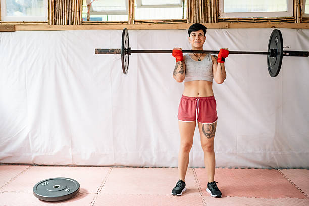 Young adult woman with tattoos and short hair lifting a barbell in a gym, focusing on strength and fitness