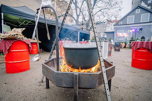 Large black cauldron suspended over an open fire, surrounded by red barrels and tents, creating a warm atmosphere for outdoor cooking and gathering experiences