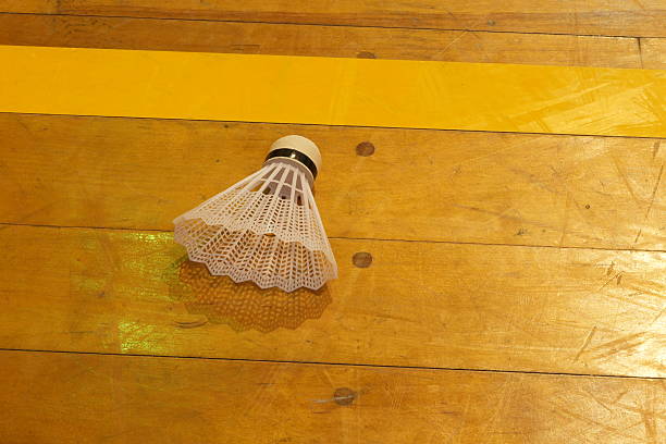 A white badminton racket is laying on a wooden floor. The racket is upside down and the strings are visible. Concept of relaxation and leisure, as the badminton racket is not in use