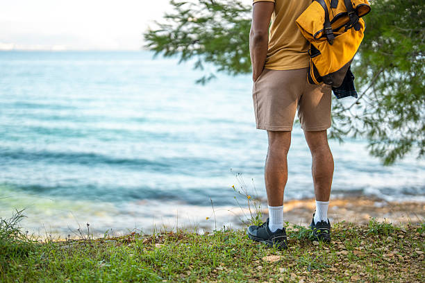 Hiker wearing khaki shorts and yellow backpack pauses on coastal trail, surrounded by green foliage while overlooking tranquil turquoise waters.