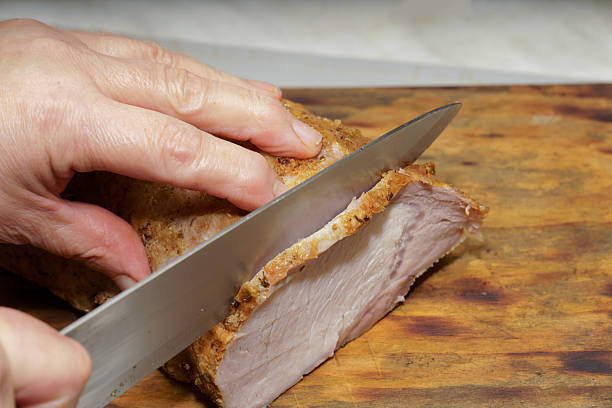 Hand slicing a piece of cooked roast meat with a knife on a wooden cutting board, preparing food in a kitchen