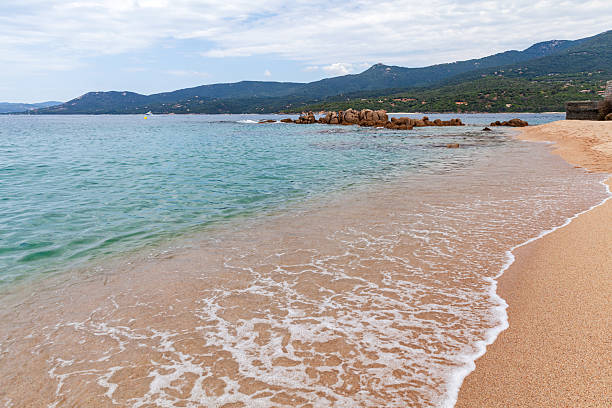 A serene coastal view featuring sea waters, sandy shoreline, rocky outcrops, and a backdrop of mountains against a partially cloudy sky, evoking a sense of tranquility and natural beauty. Corsica