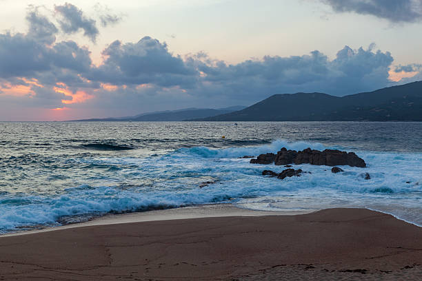 Dramatic view of shoreline with rocky formations, gently lapping waves, and a serene evening sky showcasing a vibrant sunset. Landscape of Propriano, Corsica