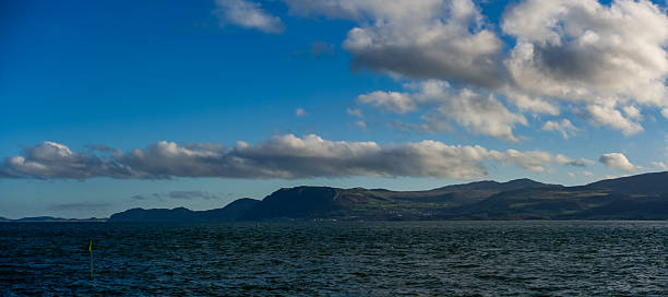 Open water stretches across the Menai Strait toward low hills on the far shore as layered clouds drift through clear blue sky creating a calm coastal atmosphere