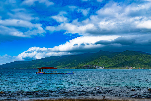 This landscape image captures a solitary floating wooden structure (likely a fishing platform or raft) anchored in the churning blue waters of a bay or ocean. The background features a majestic range of verdant green mountains stretching across the horizon, with a cluster of white buildings—possibly a coastal resort—visible along the shoreline. Above, the sky is filled with dramatic, billowing white and grey clouds set against a deep blue expanse, creating a vibrant and atmospheric coastal scene.