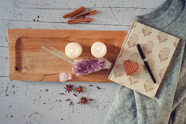 An inviting flat lay with candles on a wooden tray, amethyst and rose quartz crystals, a notebook with wooden heart, pen, and a folded grey sweater. Surrounded by cinnamon and star anise, this image evokes comfort and spiritual grounding for New Year’s reflections. Copy space included.