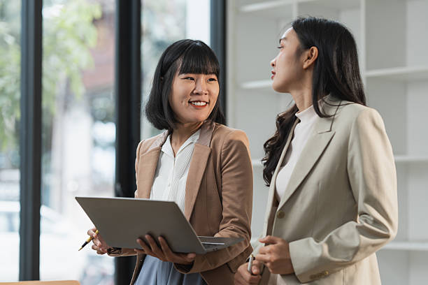 istock Two asian businesswomen engaged in a productive discussion while holding a laptop, highlighting teamwork and collaboration within a modern office setting filled with natural light 2253228457