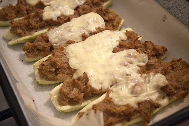 Homemade stuffed zucchini baked with melted cheese, placed on baking paper in a tray. Close-up view of cooked meal.