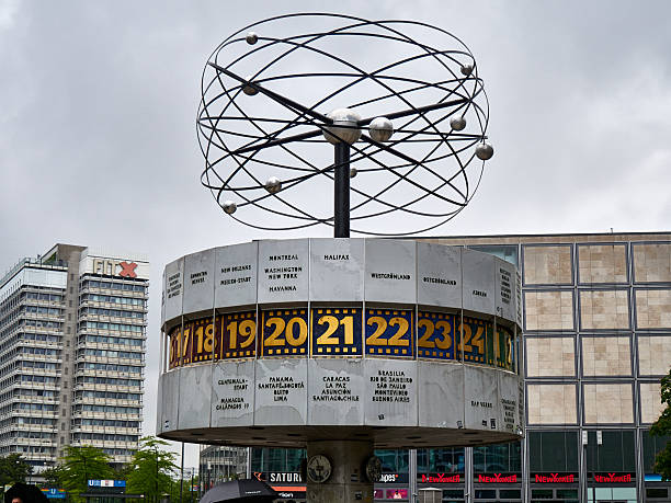A wide shot of the iconic World Time Clock (Weltzeituhr) on a rainy day at Alexanderplatz, Berlin. The photo shows the socialist-era landmark with people carrying umbrellas, a construction crane in the background, and modern retail buildings. A famous meeting point and tourist attraction in the German capital.