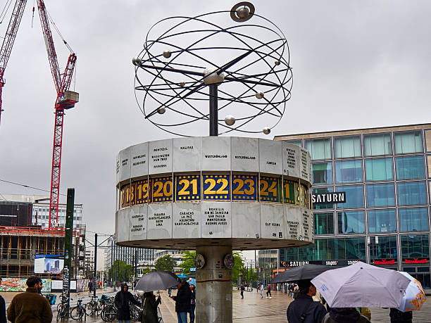 A wide shot of the iconic World Time Clock (Weltzeituhr) on a rainy day at Alexanderplatz, Berlin. The photo shows the socialist-era landmark with people carrying umbrellas, a construction crane in the background, and modern retail buildings. A famous meeting point and tourist attraction in the German capital.