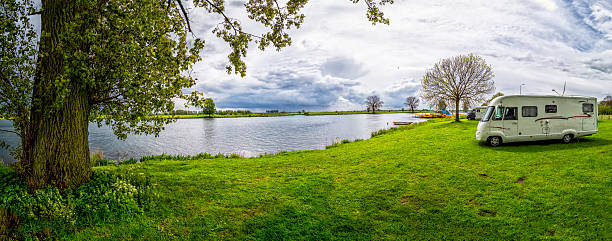 Motorhome parked on green grass by a lake with trees under a cloudy sky
