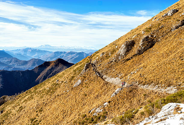Two hikers walk along a narrow mountain trail crossing a steep grassy slope in the Italian Alps. The landscape shows golden alpine meadows, rocky limestone outcrops, and layered mountain ridges, with the Monte Rosa massif visible on the horizon. The scene is captured in autumn under a clear blue sky and represents hiking, outdoor adventure, alpine tourism, and the natural beauty of Northern Italy.