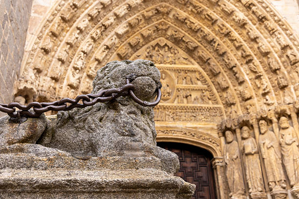 A lion statue is chained to a stone pillar in Avila. The stone pillar is part of a building with a large archway. The lion statue is chained to the pillar to prevent it from being moved or stolen