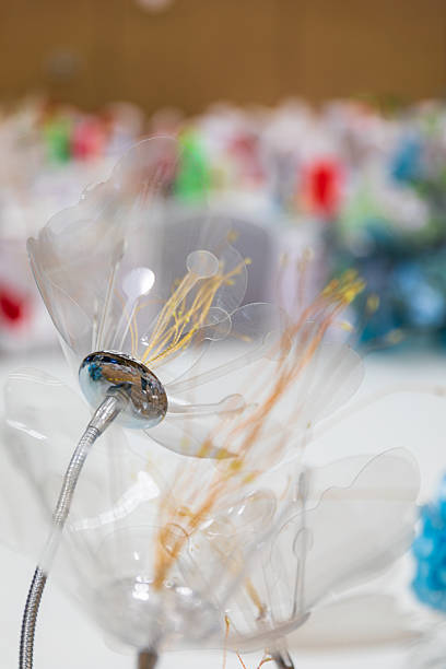 A close-up of clear tall candle holders and lush floral arrangements in yellow and white that run along the table. In the blurred background are round banquet tables with white chair covers and gift bags, warm lighting producing a soft glow. The shallow depth of field emphasizes the texture and colors of the centerpiece while conveying an elegant, festive mood.