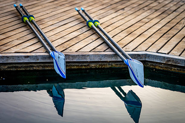 Rowing oars rest on a wooden dock by a calm lake, reflecting in the water. The scene evokes themes of, captured with a close-up photo, teamwork and sports training, ideal for a double scull setting. The serene environment suggests preparation or rest between intense rowing sessions.