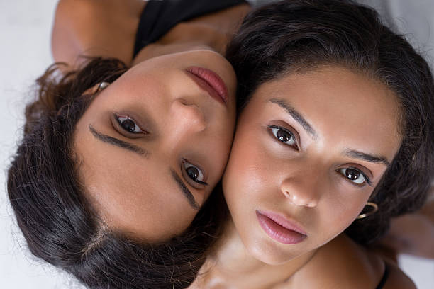 Close up portrait of twin sisters lying together and looking at the camera. Natural light, emotional bond, sisterhood, beauty and intimacy concept for lifestyle, wellness and advertising use.