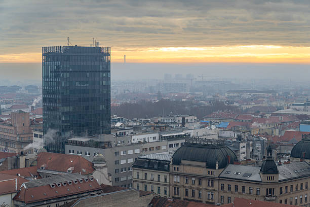 View of a city combining modern glass skyscraper and historic buildings with detailed rooftops under a cloudy sky during dusk. The composition features mixed architectural styles with a distant horizon and soft natural light
