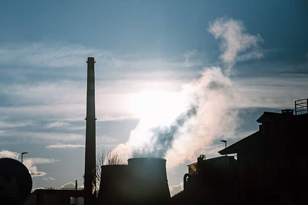Dark silhouette of a factory releasing smoke against a bright sky with the sun. Concept of industrialization, pollution, environmental impact, and dramatic landscape.