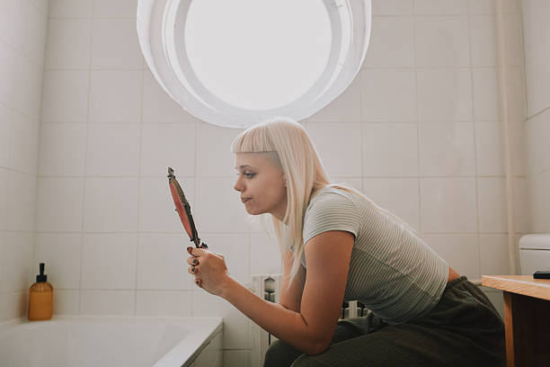 A young hipster girl sits on the toilet in her bathroom, looking at herself in the hand mirror under a round window. A calm, reflective moment that captures her morning routine and personal care.