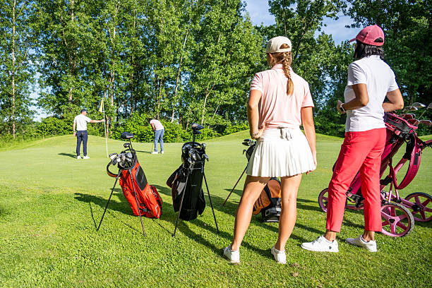 Diverse male and female golfers enjoying a day on the green, standing with golf trolleys and clubs under a clear sky, surrounded by lush trees and vibrant grass.