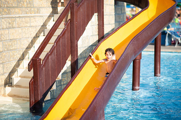 Preschool age boy is enjoying summer vacation, sliding down a yellow water slide into a blue swimming pool.