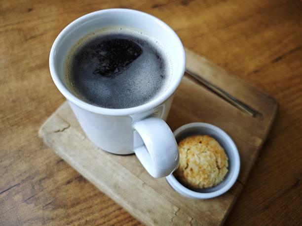 Minimalist food and beverage photograph featuring a cup of freshly brewed black coffee with crema, accompanied by a small cookie. Set on a rustic wooden board and table, creating a warm and cozy atmosphere. Suitable for concepts such as morning routine, café culture, comfort, and lifestyle.