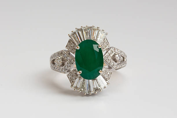 A gold ring featuring an oval cut green emerald surrounded by baguette cut diamonds on a clean white background
