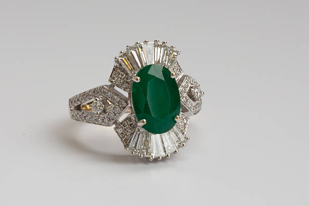 A gold ring featuring an oval cut green emerald surrounded by baguette cut diamonds on a clean white background