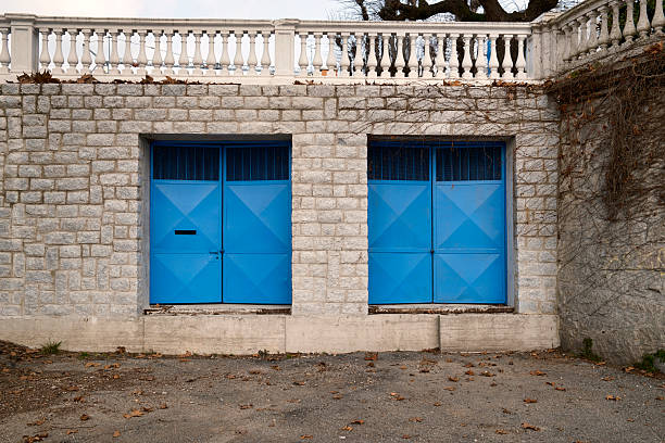 Two bright blue metal doors set into a light stone wall, creating a strong symmetrical architectural composition. Minimalist urban exterior with geometric patterns, neutral tones, and copy space, suitable for concepts such as symmetry, balance, security, simplicity, and urban design.