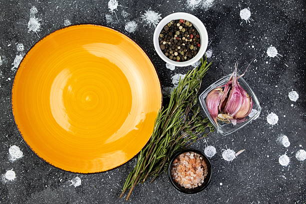 A yellow plate sits on a kitchen counter surrounded by fresh herbs, garlic, assorted peppercorns, and sea salt, ready for meal preparation.