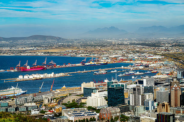 A sweeping aerial view of Cape Town’s harbor with cranes, ships, and a bustling skyline framed by Table Mountain, the Twelve Apostles, and Camps Bay in the distance.